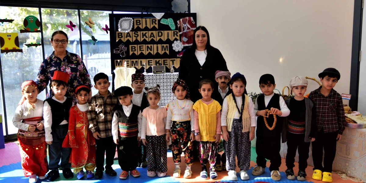 Yerli Malı Haftası etkinliği kapsamında geleneksel kıyafetler giymiş bir grup anaokulu öğrencisi, iki öğretmenleriyle birlikte yan yana duruyor. Çocukların arkasındaki panoda 
