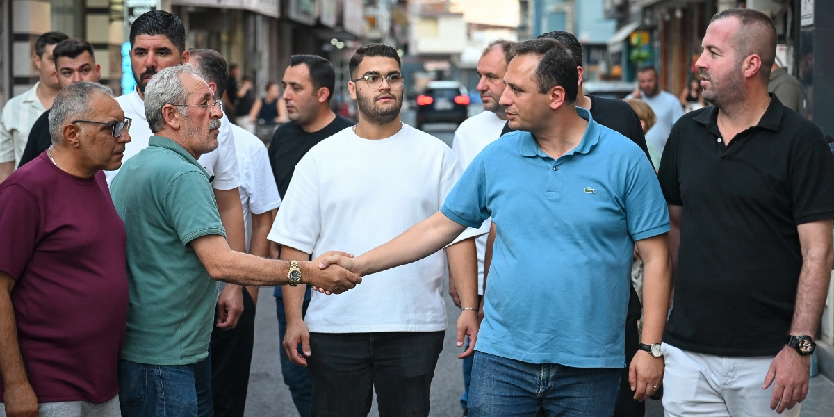 Mavi polo yaka tişört giyen Başkan Eşki, Çamdibi'ndeki bir sokakta esnafla tokalaşıyor. Çevresinde çeşitli yaş gruplarından erkeklerin bulunduğu bir kalabalık eşlik ediyor.