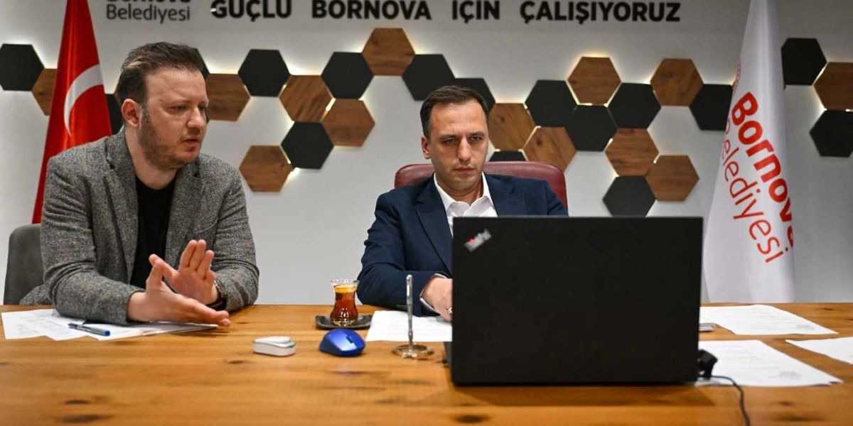 Bornova Belediyesi ofisinde, ahşap bir masa başında oturan iki erkek, önlerindeki dizüstü bilgisayara bakarak çevrimiçi bir toplantı gerçekleştiriyor. Soldaki kişi gri ceketli, sağdaki kişi ise lacivert takım elbiseli. Arka planda 