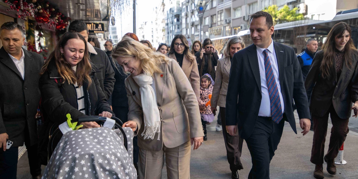 CHP Kadın Kolları Başkanı Asu Kaya ve Bornova Belediye Başkanı Ömer Eşki, beraberindeki heyetle birlikte şehir merkezinde yürürken, Asu Kaya bebek arabasındaki bir bebeği severken görülüyor. Arka planda dükkanların ve yayaların bulunduğu hareketli bir cadde atmosferi yer alıyor.