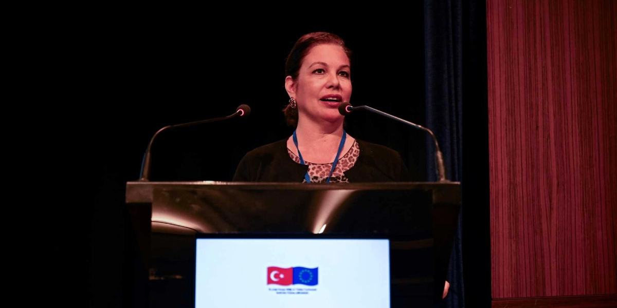Karanlık bir sahnede, kürsüde konuşma yapan orta yaşlı bir kadın. Kürsünün ön yüzündeki ekranda yan yana Türkiye ve Avrupa Birliği bayrakları yer alıyor. Kadın, siyah bir ceket ve leopar desenli bir bluz giymiş, boynunda mavi bir yaka kartı asılı.