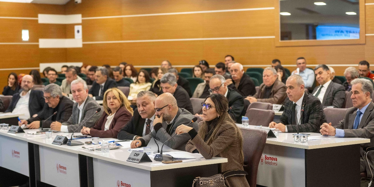 Bornova Belediyesi meclis salonunda düzenlenen toplantıda, masalarda oturan ve önlerinde mikrofonlar bulunan belediye meclis üyeleri yer alıyor. Ön planda, konuşma yapan gözlüklü bir kadın ve yanında oturan diğer meclis üyeleri görülürken, arka planda toplantıya katılan kalabalık bir grup izleyici bulunuyor. Masaların üzerinde Bornova Belediyesi logoları ve meclis üyelerinin unvanlarını belirten isimlikler yer alıyor.
