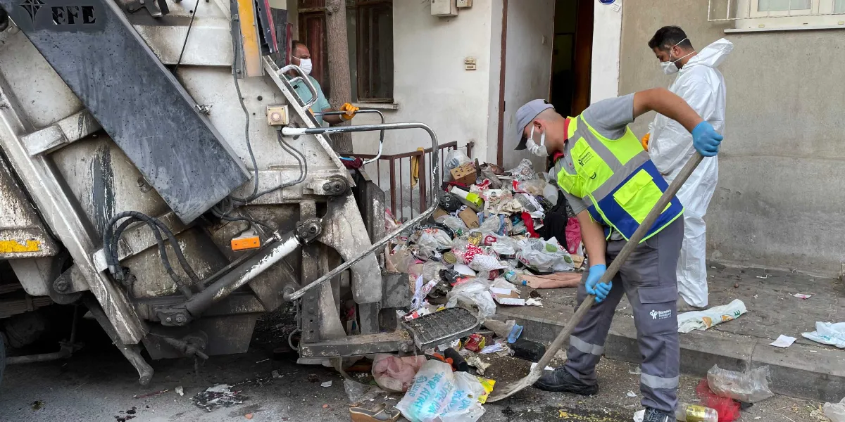 Bornova Belediyesi temizlik ekipleri, bir apartman girişinde birikmiş yoğun çöp yığınını kürek ve süpürgelerle temizleyerek çöp kamyonuna yüklüyor. Maske ve eldiven takan görevlilerden biri, üzerinde Bornova Belediyesi logosu bulunan gri üniforma ve fosforlu yelek giyerken, diğer görevli beyaz koruyucu tulum içinde çalışıyor.