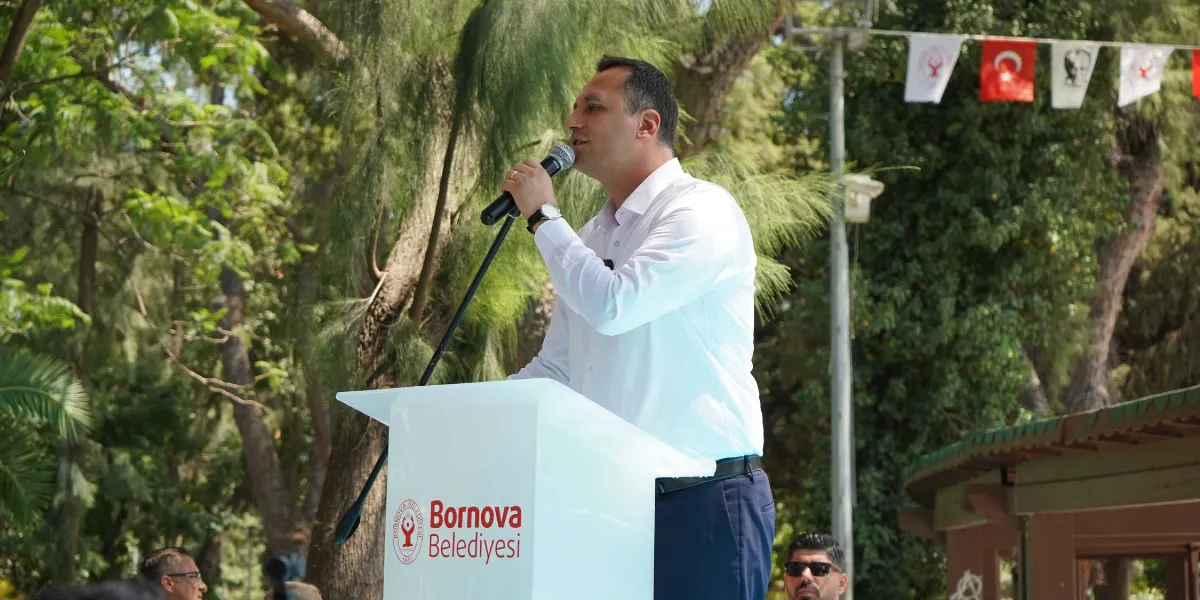 Açık havada, ağaçlık bir alanda kurulan kürsüde konuşma yapan beyaz gömlekli bir erkek. Kürsünün üzerinde Bornova Belediyesi logosu ve yazısı yer alıyor. Arka planda ise Türk bayrakları ve çeşitli flamalar asılı.