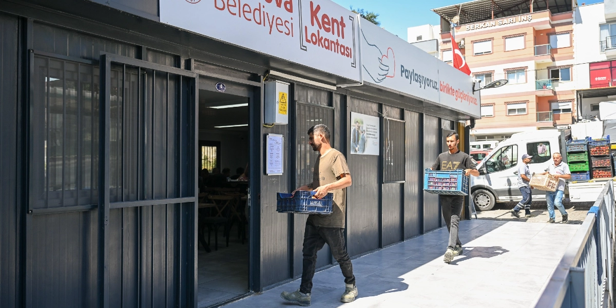Bornova Belediyesi Kent Lokantası yazılı tabelası bulunan prefabrik yapının önünde, çalışanlar ellerinde taşıdıkları kasalarla içeriye malzeme taşıyor. Binanın dış cephesinde Paylaşıyoruz, birlikte güçleniyoruz sloganı yer alırken, arka planda bir kamyonetten indirme yapan diğer görevliler ve şehir dokusu görülüyor.