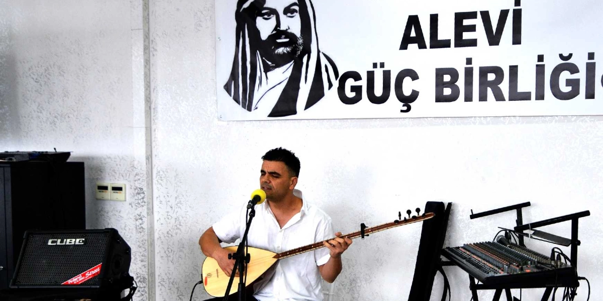 Beyaz gömlekli bir erkek, mikrofon önünde bağlama çalarak şarkı söylüyor. Arka planda üzerinde 
