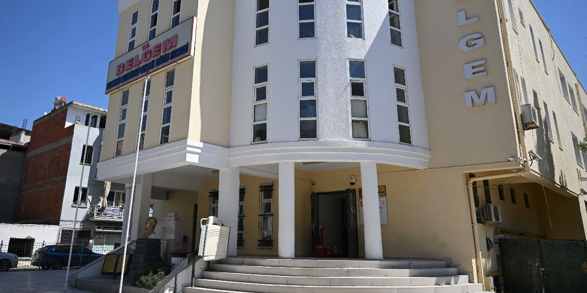 Açık sarı renkli, çok katlı ve modern mimariye sahip T.C. BELGEM Bornova Belediyesi Gençlik Eğitim Merkezi binasının dış görünüşü. Binanın girişinde beyaz sütunlar, geniş bir merdiven ve üzerinde büyük harflerle BELGEM yazan bir tabela bulunuyor. Binanın önünde küçük bir Atatürk büstü yer alıyor ve gökyüzü açık ve güneşli.