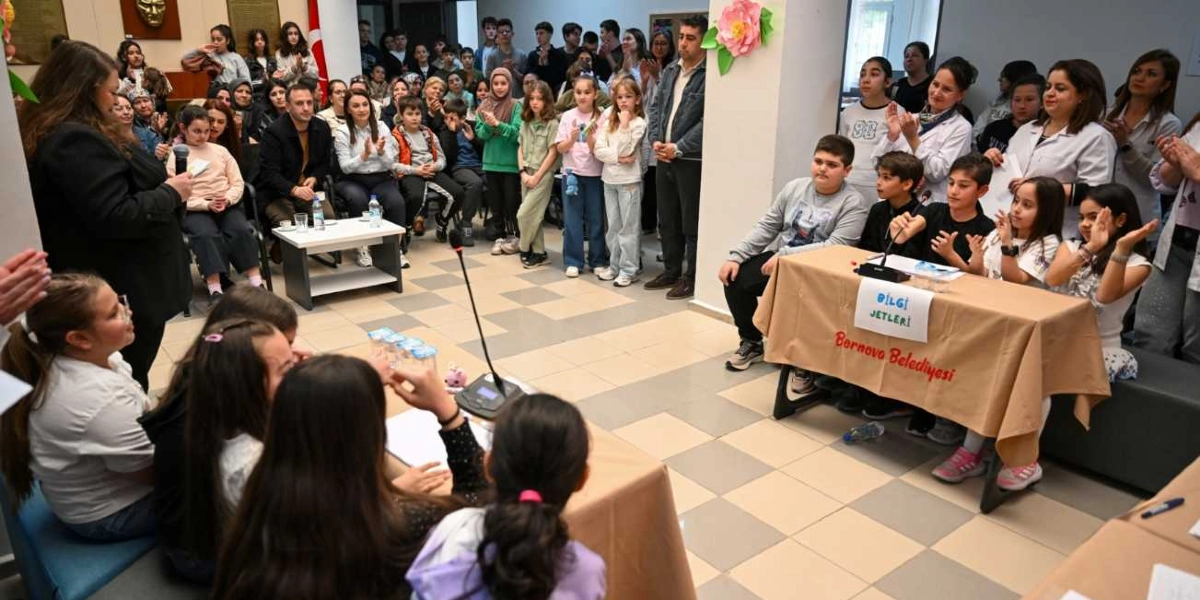 Bornova Belediyesi tarafından düzenlenen bir bilgi yarışmasında, masalarda oturan öğrenciler ve onları izleyen kalabalık bir grup yer alıyor. Sağ taraftaki masada üzerinde 