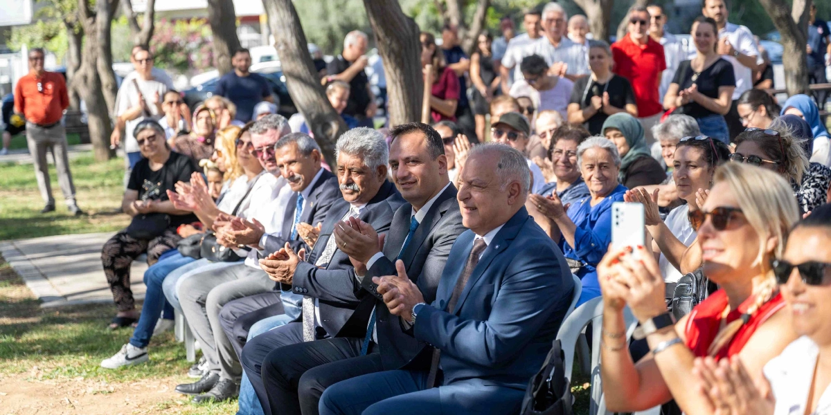 Açık havada, ağaçlık bir alanda düzenlenen etkinlikte, yan yana dizilmiş sandalyelerde oturan ve alkışlayan kalabalık bir grup insan. Ön sırada takım elbiseli üç erkek ve yanlarında çeşitli kıyafetlerle oturan kadınlar yer alıyor. En ön sağ tarafta, elindeki akıllı telefonla çekim yapan sarı saçlı bir kadın ve yanında gülümseyen başka bir kadın bulunuyor. Arka planda ise ayakta duran ve etkinliği izleyen çok sayıda katılımcı görülüyor.