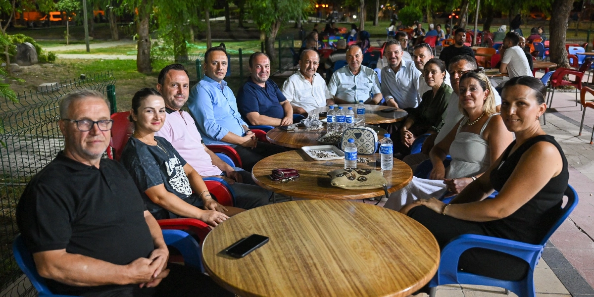Akşam saatlerinde açık hava bir park ortamında, yan yana dizilmiş yuvarlak ahşap masaların etrafında oturan kalabalık bir grup insan. Ön plandaki masada, sol tarafta gözlüklü bir bey, yanında bir kadın ve diğer katılımcılar yer alıyor. Masaların üzerinde su şişeleri ve kişisel eşyalar bulunuyor. Arka planda ise ağaçlar ve parkın diğer bölümlerinde oturan insanlar görülüyor.
