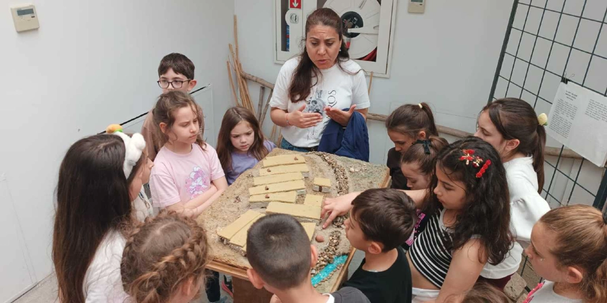 Bir kadın eğitmen, etrafına toplanmış ilkokul çağındaki bir grup çocuğa, üzerinde Taş Devri yerleşimini temsil eden maketin bulunduğu masayı anlatıyor. Çocuklar ilgiyle maketi incelerken, eğitmen elleriyle açıklayıcı hareketler yapıyor. Maketin üzerinde dikdörtgen yapılar ve küçük taşlarla çevrili bir alan yer alıyor.