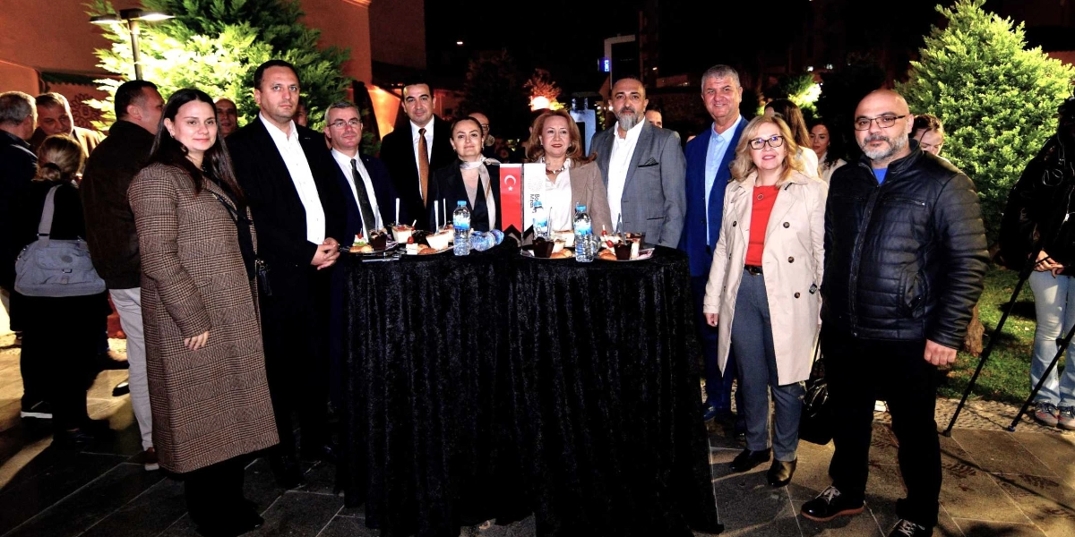 Gece vakti dış mekanda, siyah örtülü kokteyl masalarının etrafında toplanmış, resmi kıyafetli bir grup insan kameraya gülümseyerek poz veriyor. Masaların üzerinde küçük ikramlıklar ve su şişeleri bulunurken, masalardan birinin üzerinde Bornova Belediyesi'ne ait olduğu anlaşılan logolu bir flama yer alıyor. Arka planda aydınlatılmış ağaçlar ve tarihi dokuyu çağrıştıran bir çevre düzenlemesi görülüyor.