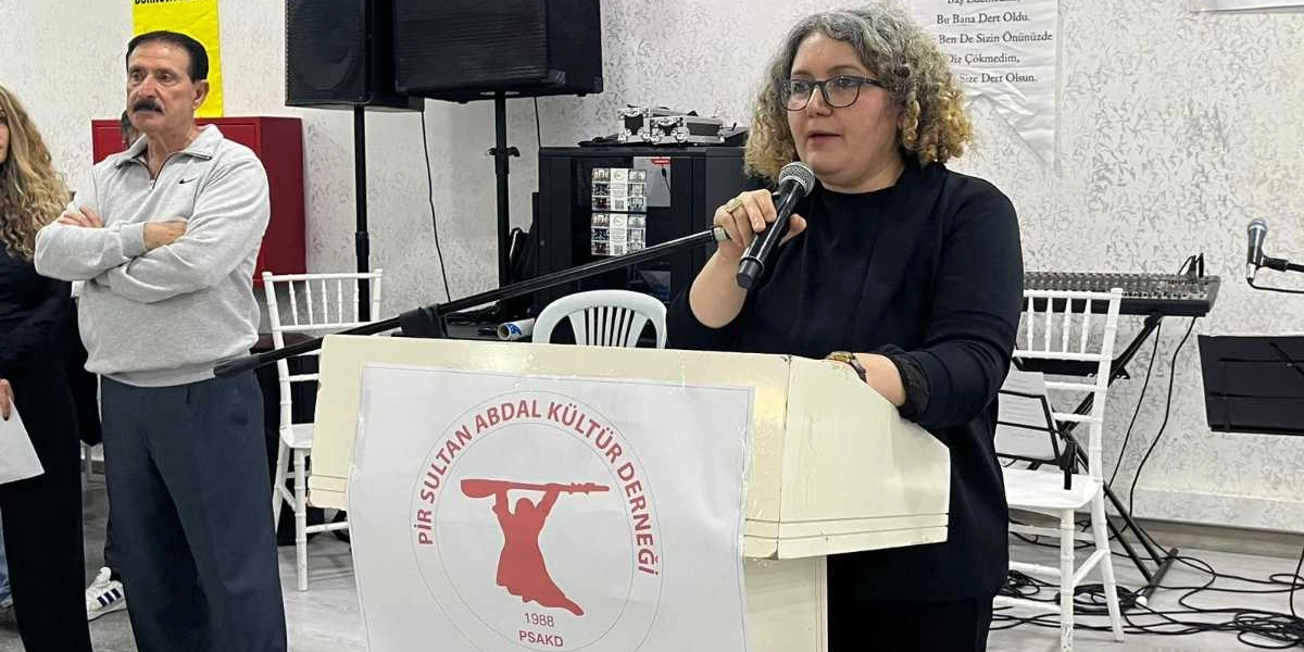 Kıvırcık saçlı ve gözlüklü bir kadın, üzerinde Pir Sultan Abdal Kültür Derneği logosu bulunan bir kürsüde mikrofonla konuşma yapıyor. Kadının sol tarafında, ellerini göğsünde kavuşturmuş şekilde duran bıyıklı bir erkek yer alıyor. Arka planda ses sistemi ekipmanları ve beyaz sandalyeler bulunuyor.