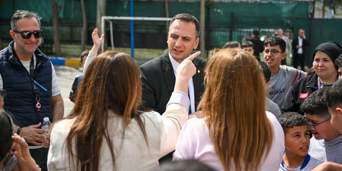 Takım elbiseli bir erkek, çevresindeki çocuklarla ve yetişkinlerle sohbet ederken eliyle bir noktayı işaret ediyor. Yanında lacivert yelekli, güneş gözlüklü bir adam ve arkası dönük, açık renkli kıyafetler giymiş kadınlar bulunuyor. Açık havada, bir okul bahçesini andıran alanda gerçekleşen bu ziyarette, çocuklar ve yetişkinler dikkatle konuşmayı dinliyor.