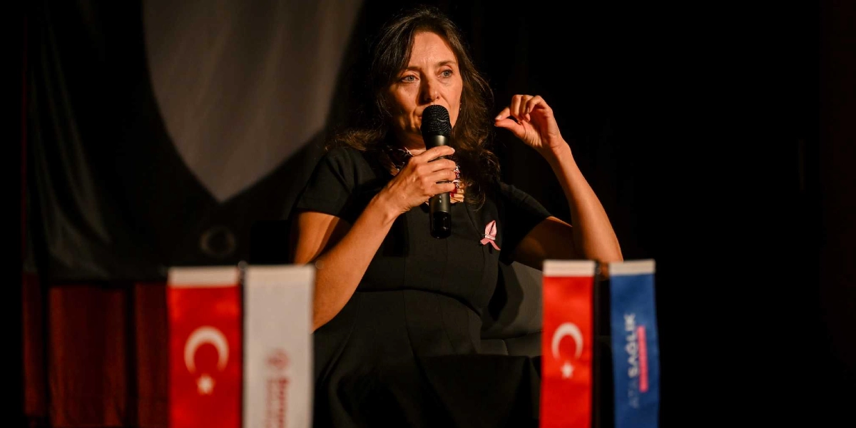 Koyu renkli bir sahnede, üzerinde pembe meme kanseri farkındalık kurdelesi bulunan siyah elbiseli bir kadın, elinde mikrofonla konuşma yapıyor. Kadının önünde, masanın üzerinde Türk bayrakları ve üzerinde 
