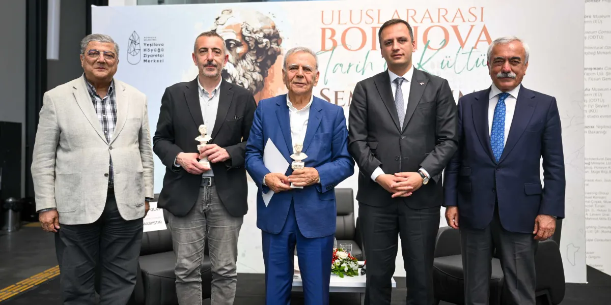 Uluslararası Bornova Tarih ve Kültür Sempozyumu sahnesinde, yan yana duran dört erkek katılımcı ve ortalarında boş bırakılmış bir koltuk yer alıyor. Katılımcılardan ikisi ellerinde küçük büstler tutuyor. Arka planda sempozyumun başlığının ve Yeşilova Höyüğü Ziyaretçi Merkezi logosunun yer aldığı bir fon bulunuyor. Boş koltuğun üzerinde 