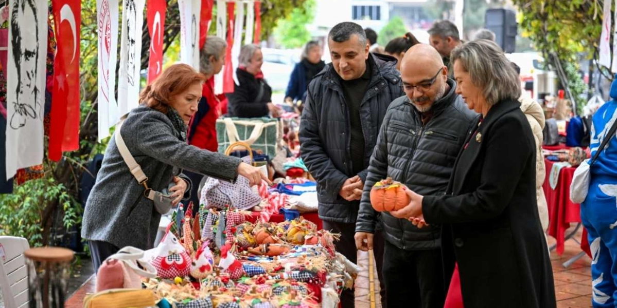 Açık havada kurulan bir hediyelik eşya pazarında, bir kadın satıcı tezgah üzerindeki el yapımı ürünleri gösterirken, yanındaki iki kişi ürünleri inceliyor. Kadın satıcı gri bir ceket giymiş, tezgahın üzerinde ise kumaştan yapılmış küçük süs eşyaları, figürler ve balkabağı şeklinde dekoratif objeler bulunuyor. Müşterilerden biri elinde turuncu renkli, balkabağı şeklinde bir süs eşyası tutuyor. Arka planda Türk bayrakları ve Atatürk silüetinin olduğu flamalar asılı, çevrede başka insanlar da alışveriş yapıyor.