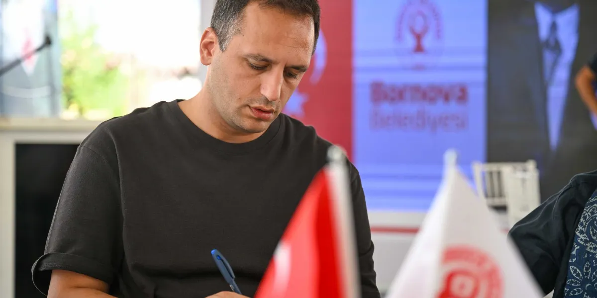 Siyah tişörtlü bir erkek, masada oturmuş elinde kalemle bir şeyler yazıyor. Önünde Türk bayrağı ve Bornova Belediyesi logolu bir masa bayrağı bulunuyor. Arka planda ise üzerinde Bornova Belediyesi yazısı ve logosu bulunan bir afiş yer alıyor.
