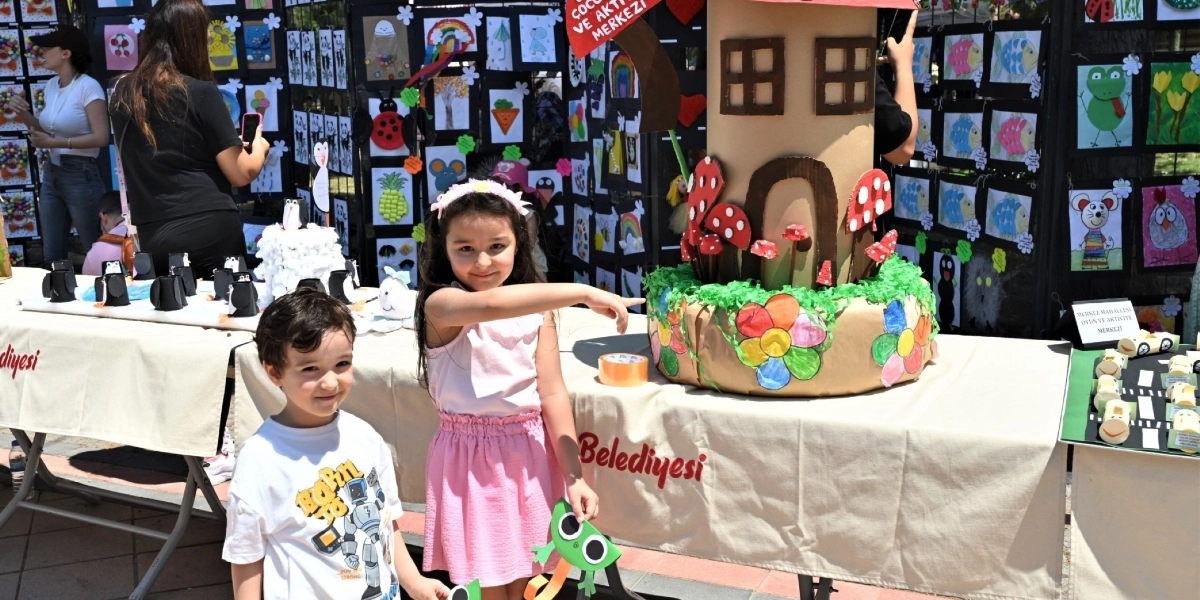 Bornova Büyükpark'ta düzenlenen çocuk sergisinde, beyaz tişörtlü bir erkek çocuk ve pembe etekli, saçında çiçekli taç olan bir kız çocuğu, el yapımı karton bir masal evi maketinin önünde poz veriyor. Kız çocuğu eliyle maketi işaret ederken, masanın üzerinde penguen figürleri ve diğer el işi çalışmalar yer alıyor. Arka planda ise panolara asılmış çok sayıda çocuk resmi ve sergiyi gezen ziyaretçiler görülüyor.