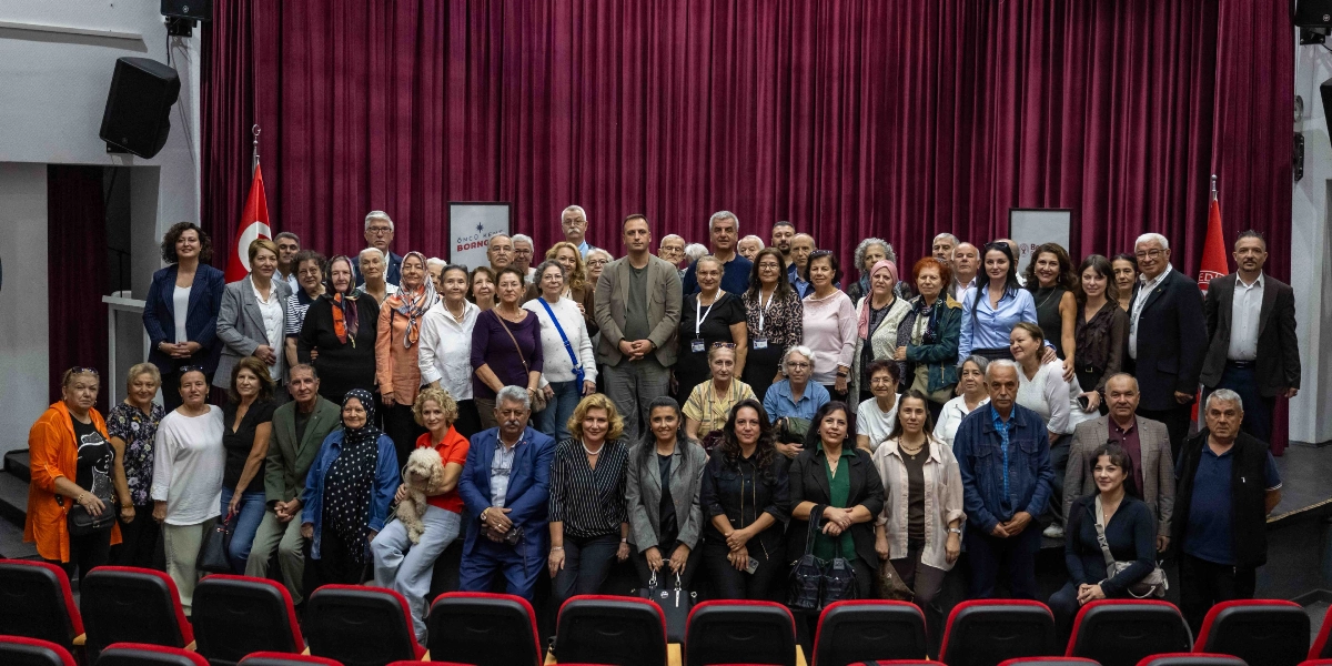Bordo renkli sahne perdelerinin önünde, farklı yaş gruplarından oluşan kalabalık bir grup insan, hatıra fotoğrafı çektirmek için yan yana dizilmiş. Grubun merkezinde Bornova Belediyesi yetkilileri olduğu düşünülen kişiler yer alırken, katılımcıların çoğu günlük kıyafetler içinde gülümseyerek kameraya bakıyor. Sahnenin ön kısmında boş koltuklar görülüyor, arka planda ise Bornova Belediyesi'ne ait logolu panolar ve Türk bayrakları bulunuyor.
