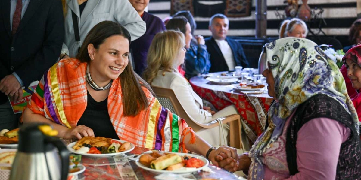 Bornova'da düzenlenen bir Anneler Günü etkinliğinde, omuzlarında renkli, geleneksel desenli bir şal bulunan genç bir kadın, karşısında oturan ve başörtüsü takan yaşlı bir kadının elini tutarak gülümsüyor. Masada kahvaltılık yiyeceklerin bulunduğu tabaklar yer alırken, arka planda etkinliğe katılan diğer insanlar ve yemek masaları görülüyor.