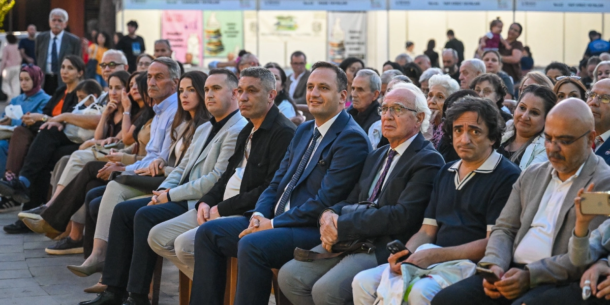 Açık havada düzenlenen bir etkinlikte, yan yana dizilmiş sandalyelerde oturan ve dikkatle sahneyi izleyen kalabalık bir izleyici grubu. Ön sırada takım elbiseli ve günlük kıyafetli çeşitli yaşlardan kadın ve erkekler yer alıyor; arka planda ise beyaz stantların bulunduğu bir fuar alanı görülüyor.