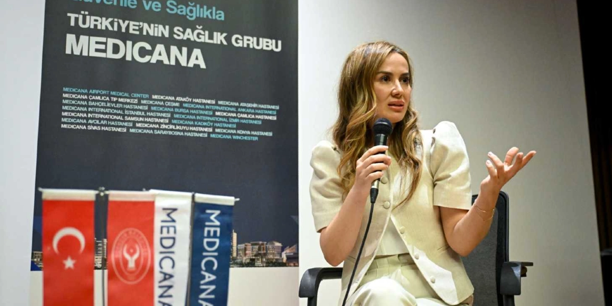 Açık renkli, düğmeli bir ceket ve pantolon giymiş bir kadın, elinde mikrofonla konuşma yapıyor. Kadının arkasında, üzerinde 