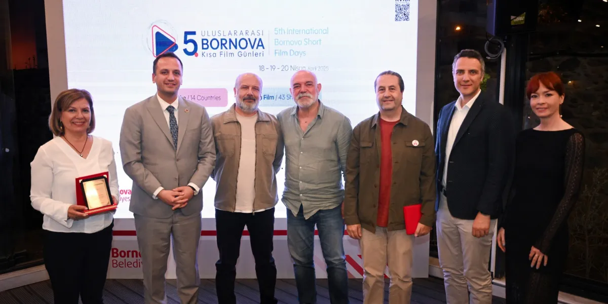 Arka planda 5. Uluslararası Bornova Kısa Film Günleri'ni tanıtan bir afişin önünde yan yana duran altı kişilik bir grup. Soldan sağa doğru; elinde bir plaket tutan beyaz bluzlu bir kadın, gri takım elbiseli bir erkek, bej ceketli sakallı bir erkek, gri gömlekli sakallı bir erkek, haki ceketli bir erkek, lacivert ceketli bir erkek ve siyah elbiseli bir kadın yer alıyor. Grup, objektife gülümseyerek poz veriyor.