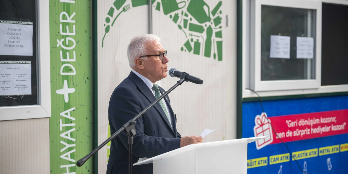Beyaz saçlı, gözlüklü ve takım elbiseli bir yetkili, Atık Bank projesinin açılışında beyaz bir kürsü arkasında mikrofonla konuşma yapıyor. Arka planda üzerinde 
