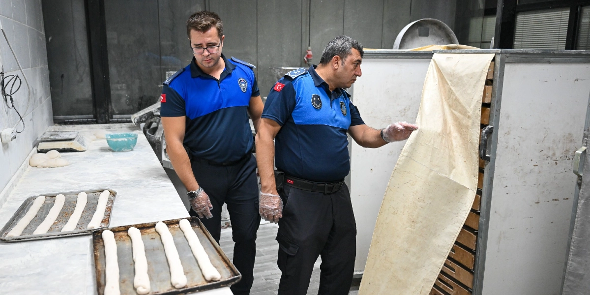 Mavi ve lacivert üniformalı iki zabıta memuru, bir fırının üretim alanında denetim yapıyor. Sağdaki memur, hamur tepsilerinin bulunduğu metal bir dolabın örtüsünü kaldırarak içini incelerken, soldaki gözlüklü memur ise tezgâh üzerindeki hamurları kontrol ediyor. Tezgâhın üzerinde pişmeye hazır hamur şeritleri ve bir kase bulunuyor.