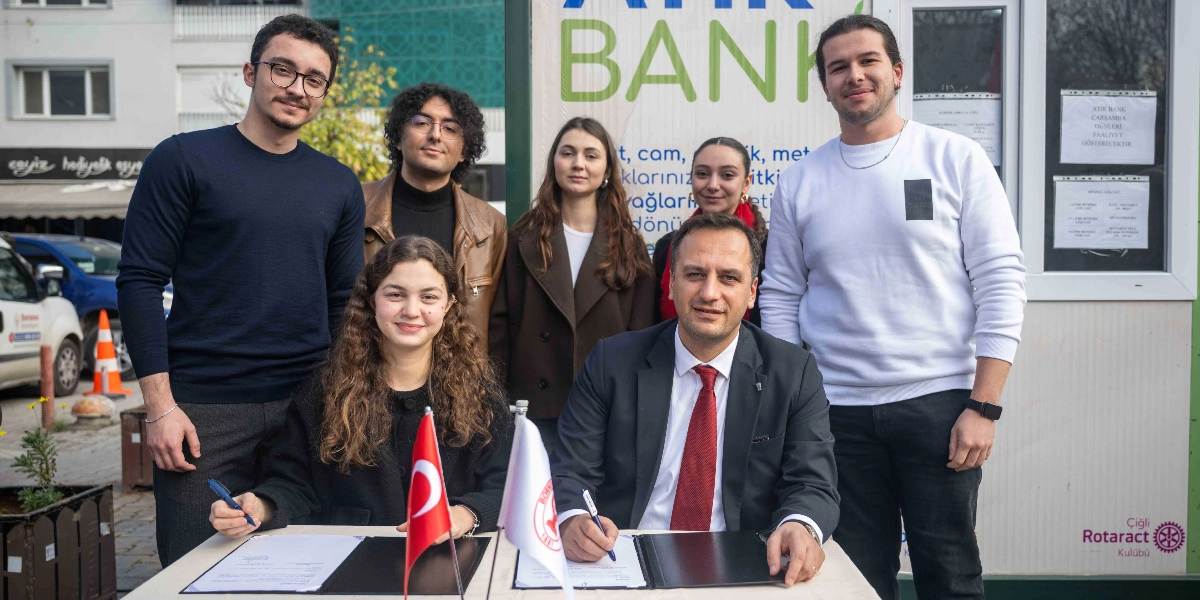 Bornova Atık Bank projesinin açılışında, bir masada karşılıklı oturarak protokol imzalayan bir kadın ve bir erkek ile arkalarında duran dört gençten oluşan bir grup. Arka planda üzerinde 