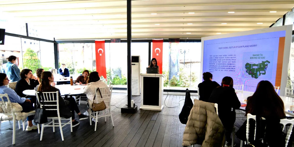 Aydınlık ve modern bir toplantı salonunda, bir konuşmacı kürsüde sunum yaparken, katılımcıların masalar etrafında oturarak dinlediği bir etkinlik. Salonun ön tarafındaki dev ekranda 
