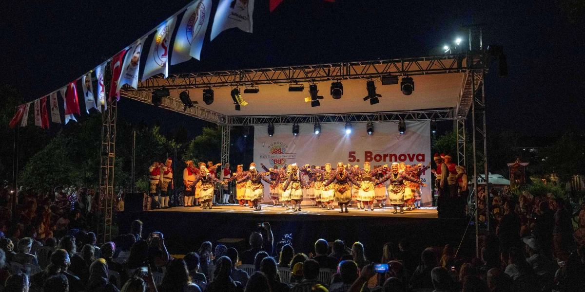 Gece vakti açık havada kurulan bir sahnede, geleneksel kıyafetler giymiş bir grup dansçı ellerinde kaşıklarla halk oyunu sergiliyor. Sahnenin arka planında 