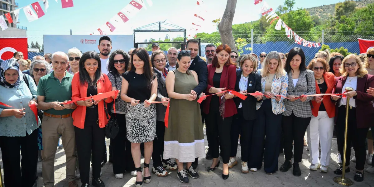 Açık havada düzenlenen bir açılış töreninde, Bornova Belediyesi logolu bir pankartın önünde yan yana dizilmiş kalabalık bir grup, ellerindeki makaslarla kırmızı bir kurdeleyi kesiyor. Çoğunluğu kadınlardan oluşan katılımcılar, günlük kıyafetler içinde gülümseyerek kurdeleyi tutuyor. Arka planda Türk bayrakları ve süslemeler yer alıyor.