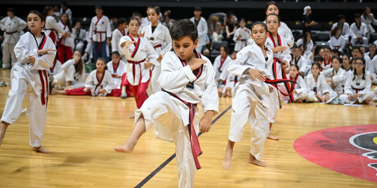 Kapalı bir spor salonunda, beyaz tekvando kıyafetleri ve renkli kuşaklar giymiş çocuklardan oluşan bir grup, antrenman veya gösteri sırasında tekvando hareketleri sergiliyor. Ön planda, kırmızı-siyah kuşaklı bir erkek çocuk tek ayağı üzerinde dengede durarak tekme atma pozisyonu alırken, yanında ve arkasında benzer hareketleri yapan diğer çocuklar yer alıyor. Arka planda ise tribünlerde oturan izleyiciler ve diğer sporcular bulunuyor.