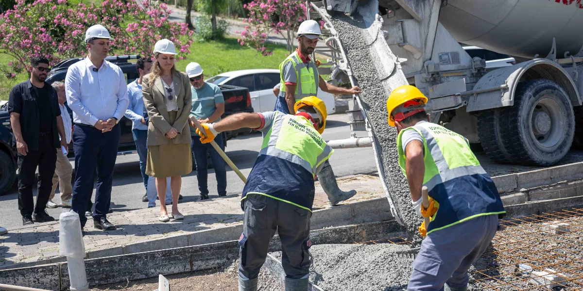 Bornova Belediyesi Fen İşleri Müdürlüğü yelekleri giymiş iki işçi, bir beton mikserinden dökülen harcı küreklerle zemine yayıyor. Çalışmaları, aralarında beyaz baretli yetkililerin de bulunduğu bir grup insan dikkatle izliyor. Arka planda yeşil ağaçlar ve park halindeki araçlar görülüyor.