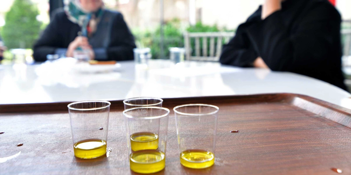 Kahverengi bir tepsi üzerinde, içinde az miktarda sarı renkli zeytinyağı bulunan üç adet şeffaf plastik tadım bardağı duruyor. Arka planda ise bulanık bir şekilde masada oturan ve sohbet eden insanlar görünüyor.
