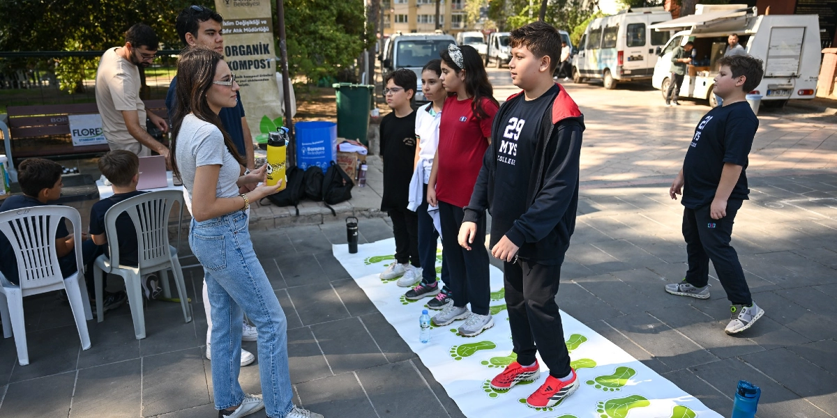 Açık havada düzenlenen bir etkinlikte, üzerinde yeşil ayak izleri bulunan uzun bir şerit üzerinde duran çocuklar ve onlara rehberlik eden bir genç kadın yer alıyor. Çocuklar sırayla şeridin üzerinde ilerlerken, arka planda Bornova Belediyesi'ne ait bilgilendirme panoları, oturma grupları ve park halindeki araçlar görülüyor.