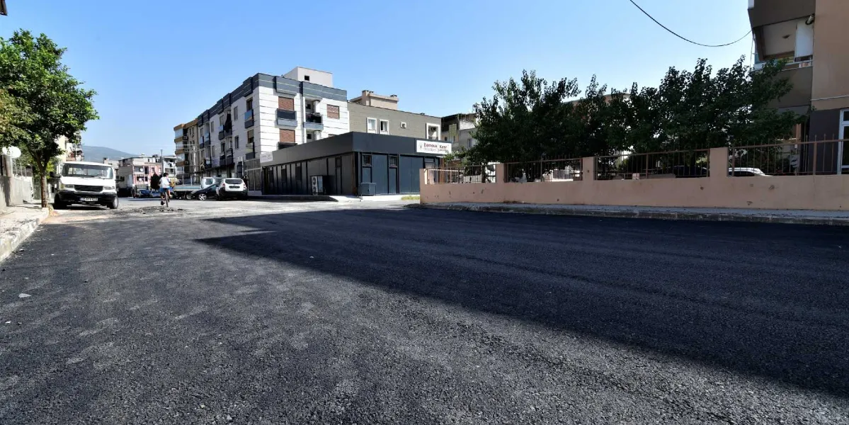 Bornova'da yeni asfaltlanmış geniş bir cadde ve arka planda çok katlı binalar ile Bornova Kent Konseyi tabelasının bulunduğu modern bir yapı görülüyor. Yolun kenarında park halindeki araçlar ve ağaçlar yer alıyor.