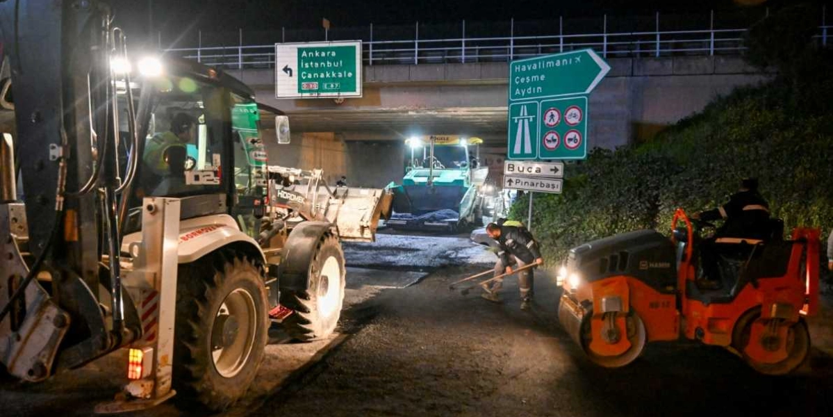 Gece saatlerinde bir üst geçidin altında devam eden yol çalışmasında, bir iş makinesi, asfalt serici ve silindir yer alıyor. İşçilerden biri elindeki aletle asfaltı düzeltirken, arka planda Ankara, İstanbul, Çanakkale, Havalimanı, Çeşme, Aydın, Buca ve Pınarbaşı yönlerini gösteren trafik tabelaları bulunuyor.