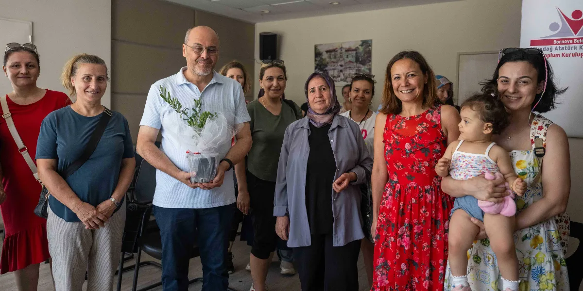 Kapalı bir mekanda yan yana duran, aralarında Bornova Belediyesi temsilcilerinin de bulunduğu bir grup kadın ve bir erkek, gıda güvenliği temalı bir etkinlik kapsamında poz veriyor. Gruptaki erkek, elinde tül ile sarılmış küçük bir zeytin fidanı tutuyor. Sağ tarafta bir kadın kucağında küçük bir çocukla yer alıyor. Arka planda Bornova Belediyesi'ne ait bir kurumun logosunun yer aldığı bir afiş ve duvarda bir tablo bulunuyor.