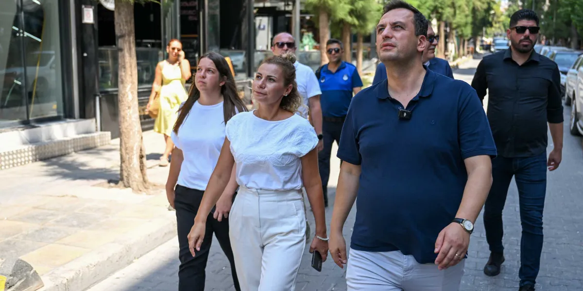 Bornova Belediye Başkanı Mustafa İduğ ve beraberindeki heyet, bir sokakta yürürken yukarıya doğru bakarak çevreyi inceliyor. Başkan İduğ lacivert polo yaka tişört ve açık renkli pantolon giymiş, yanında ise beyaz bluz ve pantolon giyen bir kadın ile diğer belediye yetkilileri yer alıyor.