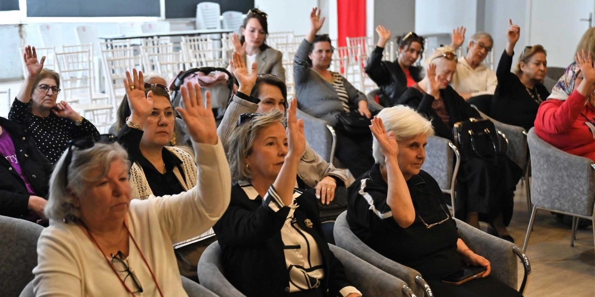 Kapalı bir salonda, gri koltuklarda oturan ve ellerini oylama yapmak için havaya kaldırmış bir grup kadın. Farklı yaş ve giyim tarzlarına sahip kadınlar, dikkatli bir şekilde ön tarafa doğru bakarak demokratik bir katılım sergiliyor.