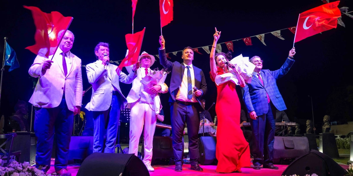 Gece vakti açık hava sahnesinde, ellerinde Türk bayrakları tutan ve mikrofonla konuşan bir grup insan yer alıyor. Soldan sağa doğru; açık renk ceketli bir beyefendi, mikrofonla konuşan gri ceketli bir kişi, elinde çiçek buketi olan beyaz takım elbiseli ve şapkalı bir beyefendi, koyu renk takım elbiseli bir beyefendi, kırmızı uzun bir elbise giymiş mikrofon tutan bir kadın ve lacivert ekose ceketli bir beyefendi bulunuyor. Arka planda ise iplere asılı küçük Türk bayrakları ve karanlık bir gökyüzü görülüyor.