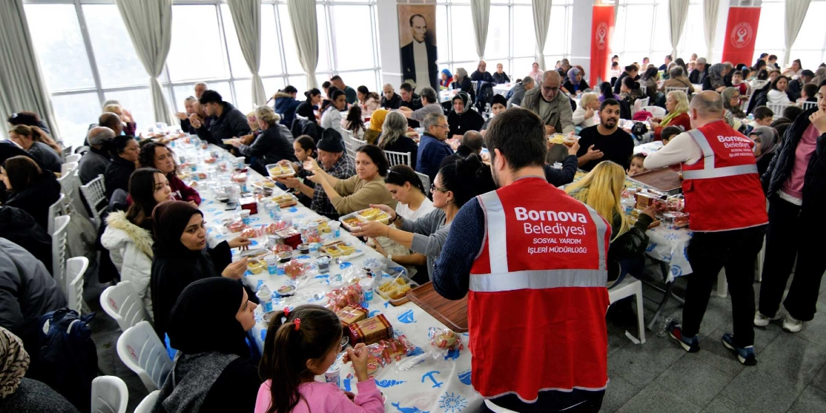 Bornova Belediyesi Sosyal Yardım İşleri Müdürlüğü görevlilerinin, kalabalık bir katılımcı grubuna yemek servisi yaptığı yılbaşı yemeği etkinliği. Uzun masalarda oturan vatandaşlara, üzerinde 