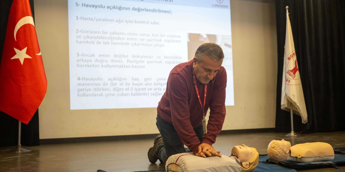 Bordo kazaklı bir erkek, sahnede yerdeki ilk yardım mankeni üzerinde kalp masajı uygulaması yaparken görülüyor. Arkasındaki projeksiyon perdesinde 