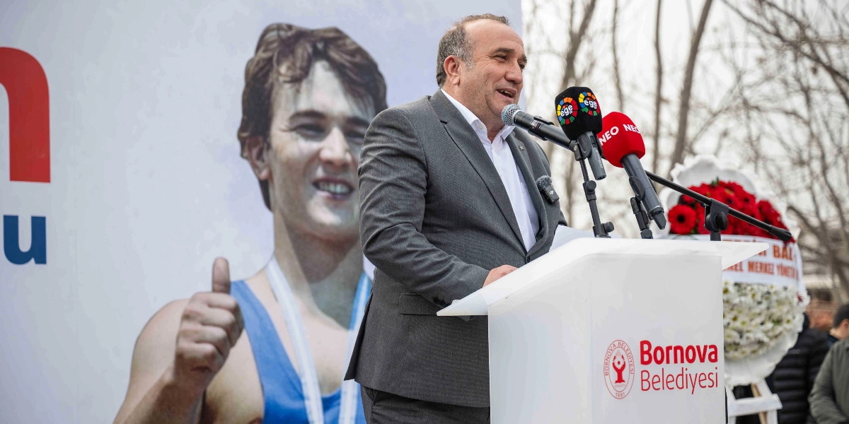 Bornova Belediye Başkanı, üzerinde Bornova Belediyesi logosu bulunan bir kürsüde konuşma yapıyor. Arka planda, halterci Naim Süleymanoğlu'nun gençlik yıllarına ait, baş parmağıyla 