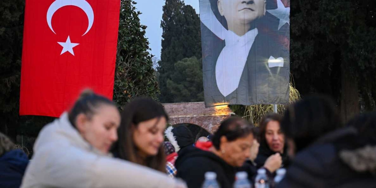 Açık havada düzenlenen bir iftar yemeğinde, masalarda oturan insanlar ve arka planda asılı duran büyük bir Türk bayrağı ile Mustafa Kemal Atatürk posteri görülüyor. Akşam saatlerinde çekilen karede, katılımcılar yemeklerini yerken ön planda odak dışı kalmış kişiler yer alıyor.