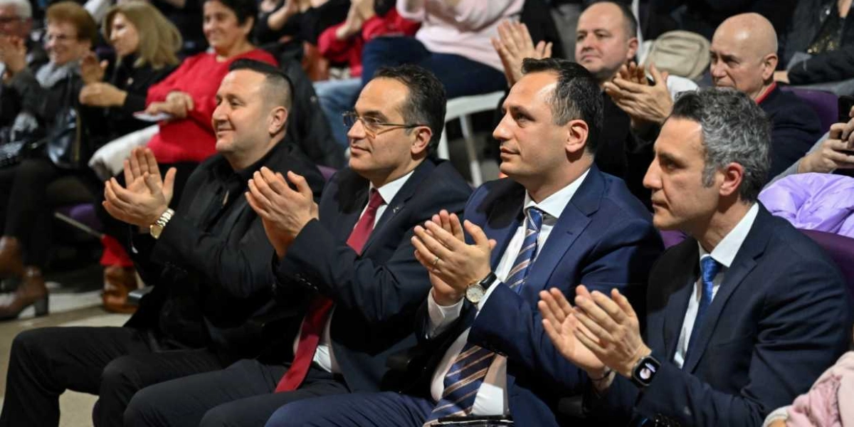 Kapalı bir salonda, takım elbiseli dört erkek yan yana oturmuş, önlerindeki sahneyi izleyerek alkışlıyor. Arka planda ise etkinliği takip eden diğer izleyiciler yer alıyor.