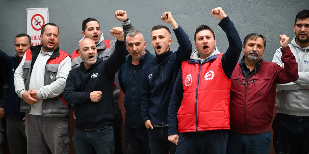 Bornova Belediye Başkanı Ömer Eşki ve bir grup işçi, gri bir duvarın önünde yan yana durarak yumruklarını havaya kaldırmış ve slogan atar bir ifadeyle poz veriyor. Ömer Eşki, üzerinde kırmızı bir yelek ve DİSK amblemiyle grubun ön sıralarında yer alıyor. İşçilerin birçoğu Bornova Belediyesi logolu kıyafetler giyiyor ve arka planda duvarda 
